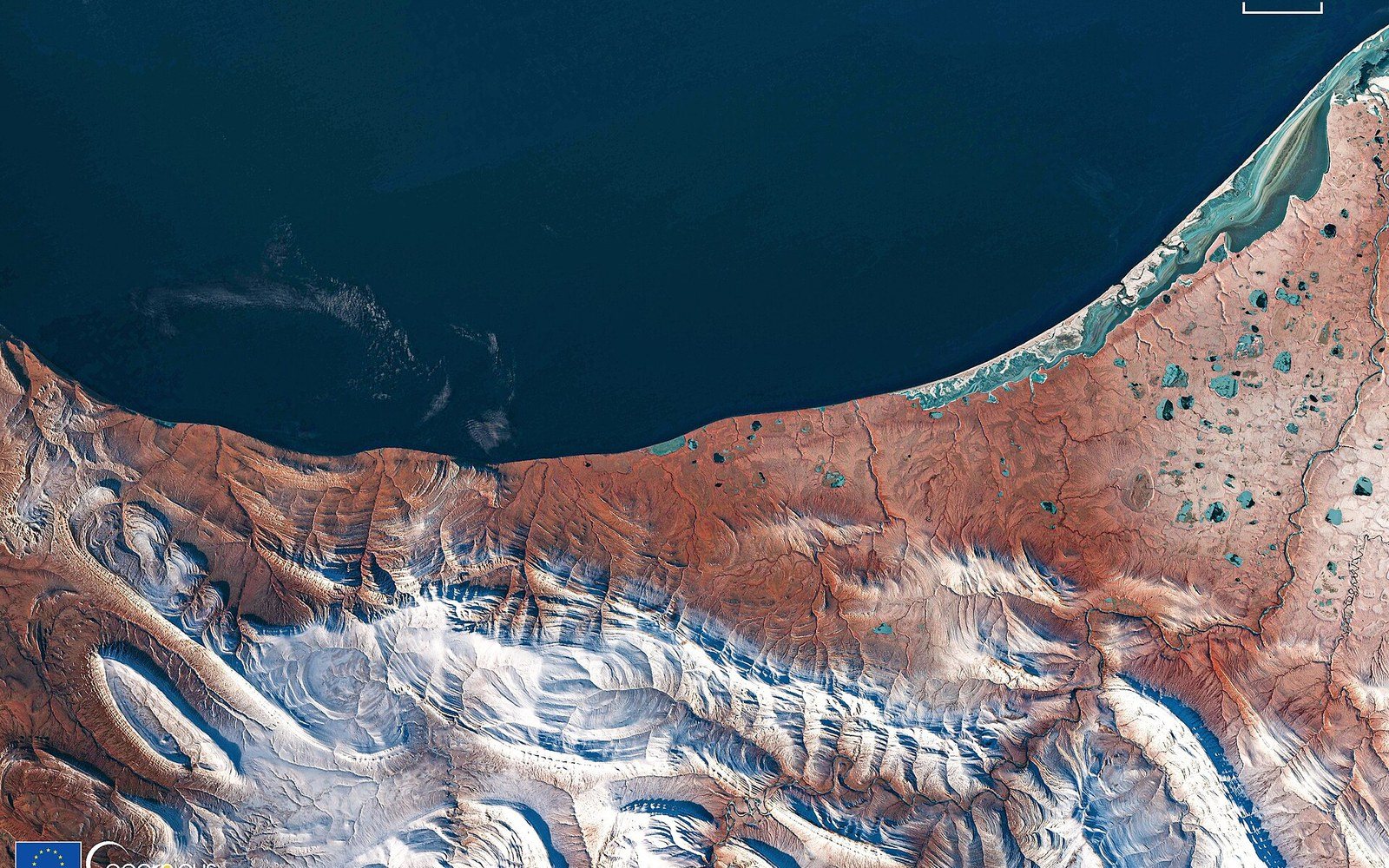 Satellite view of a rugged coastline meeting dark open water, with snow-streaked mountains inland